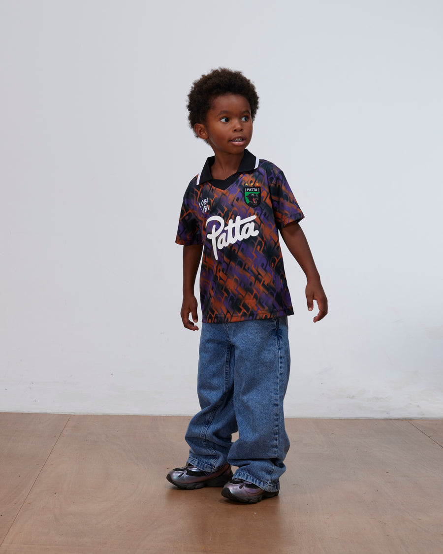 Young boy wearing the Patta Kids All Over P Football Jersey in pansy purple and orange with jeans.
